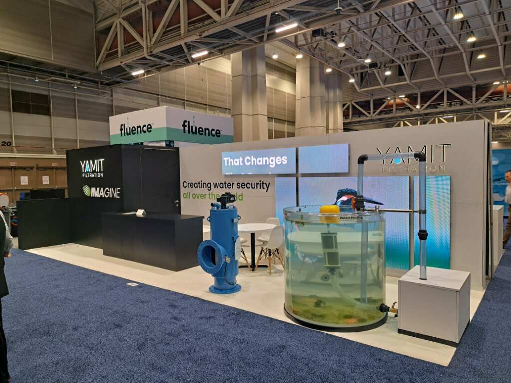 A trade show booth featuring Yamit Filtration and Fluence Corporation. The booth showcases water security technology, with a central display including a large, clear cylindrical water tank. A blue water valve model and informational signs are also present.