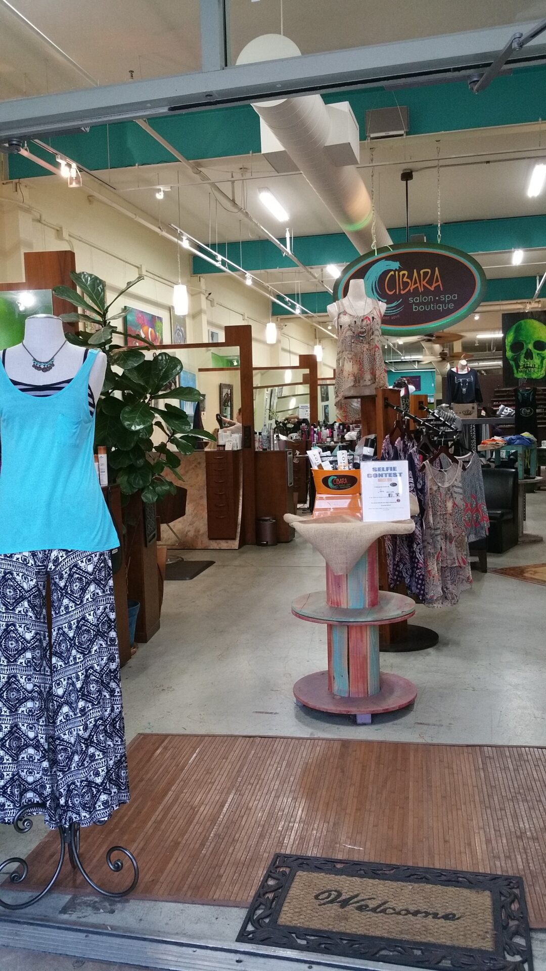 Interior view of a fashion boutique named Cibara, showcasing a mannequin wearing a blue top and patterned pants near the entrance. Various clothing items are displayed on racks and shelves throughout the store. A "Welcome" mat is placed at the entrance.