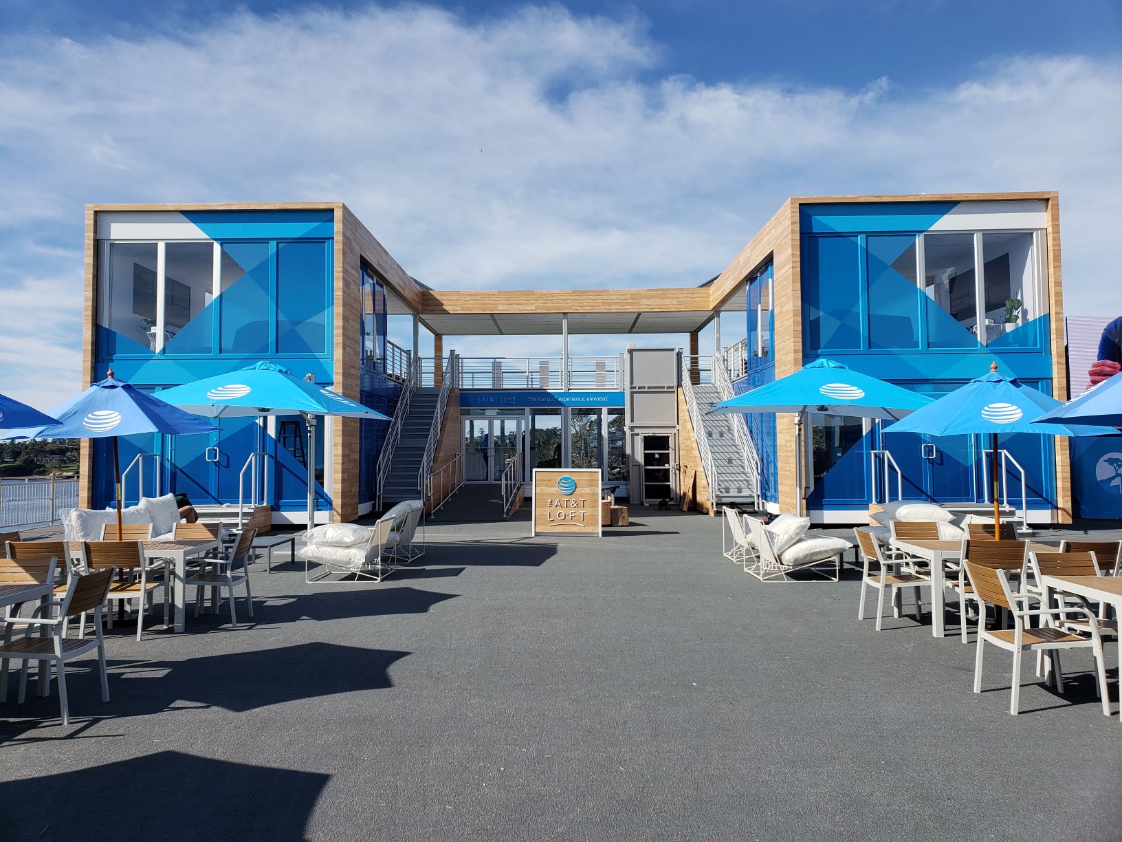 Outdoor seating area with chairs, tables, and blue umbrellas arranged on a paved surface. There are two blue and wood-paneled buildings with large windows and staircases leading up to a second level. A blue sky and water body are visible in the background.