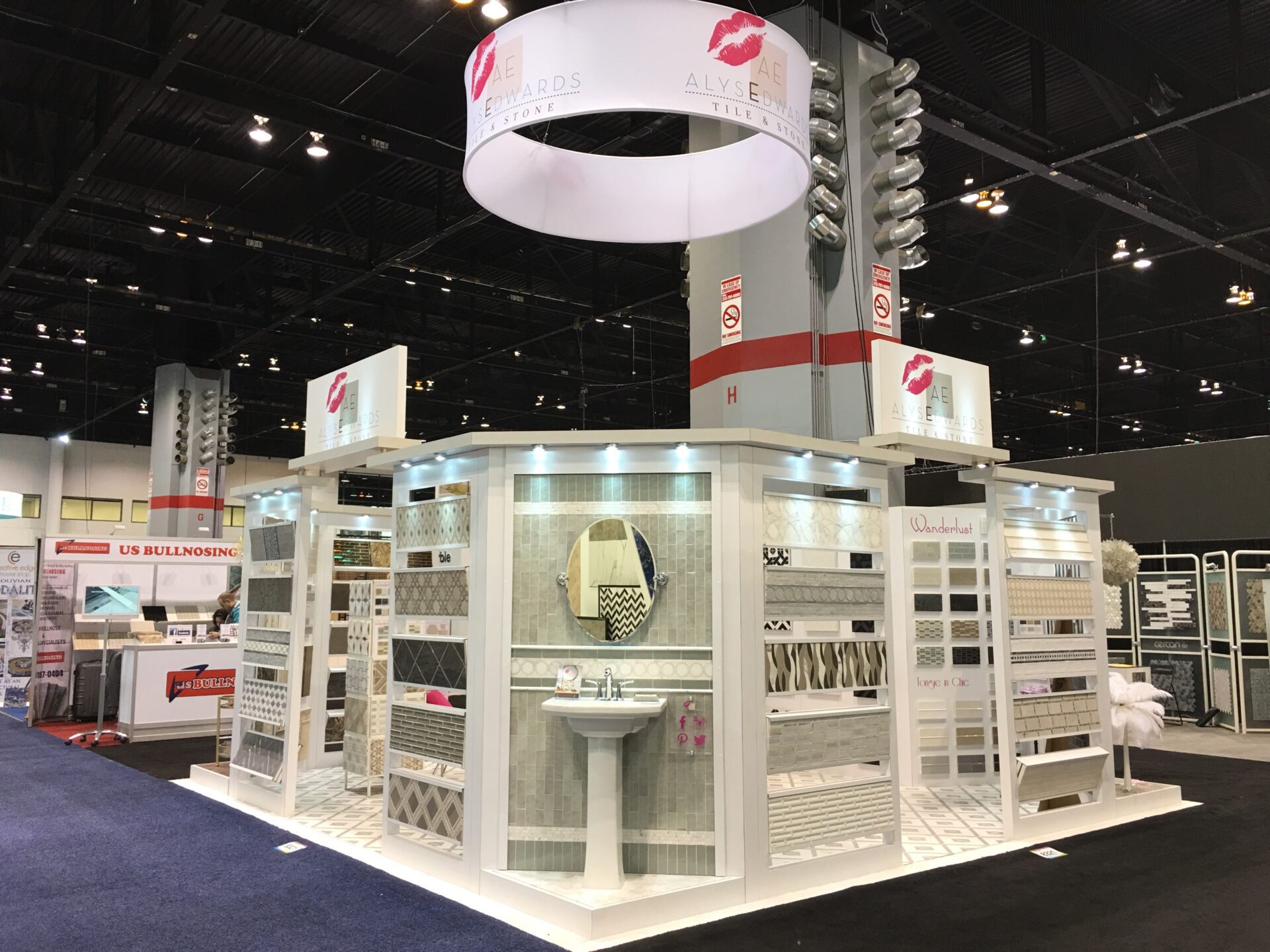 A trade show booth featuring a variety of tile displays. The booth has white walls, with sample tiles of various patterns and colors displayed. A circular sign with lips logo and the words "Awards" and "Alyse" hangs above. The booth is well-lit and organized.