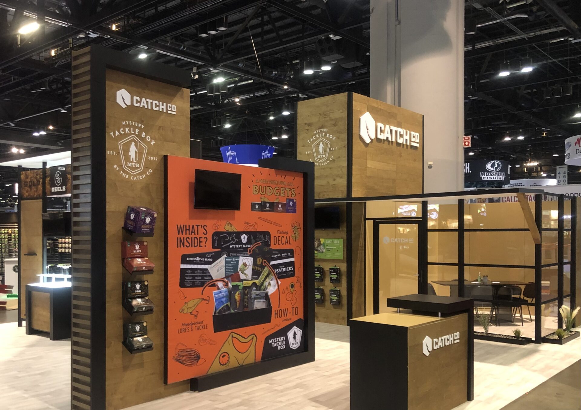 A trade show booth for Catch Co. features a large wooden structure with branding and logos. The booth includes a display screen showing "Budget's Buds" with various product illustrations, sample packs, and seating areas. The background displays other exhibition booths.