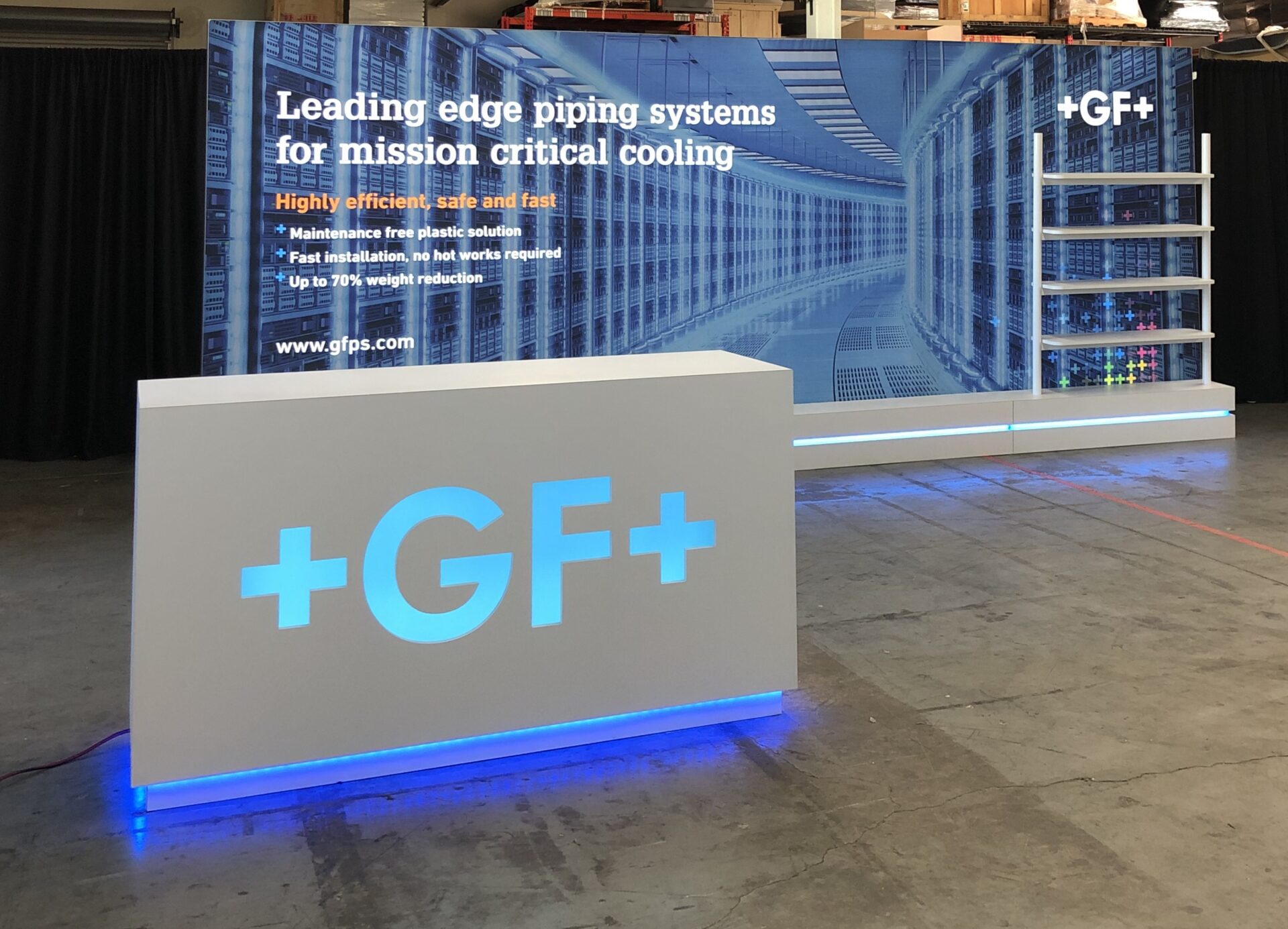 A trade show booth with a white reception counter labeled "+GF+" in blue. Behind it is a backdrop displaying text about piping systems for mission-critical cooling, highlighting features like high efficiency, easy installation, and weight reduction.