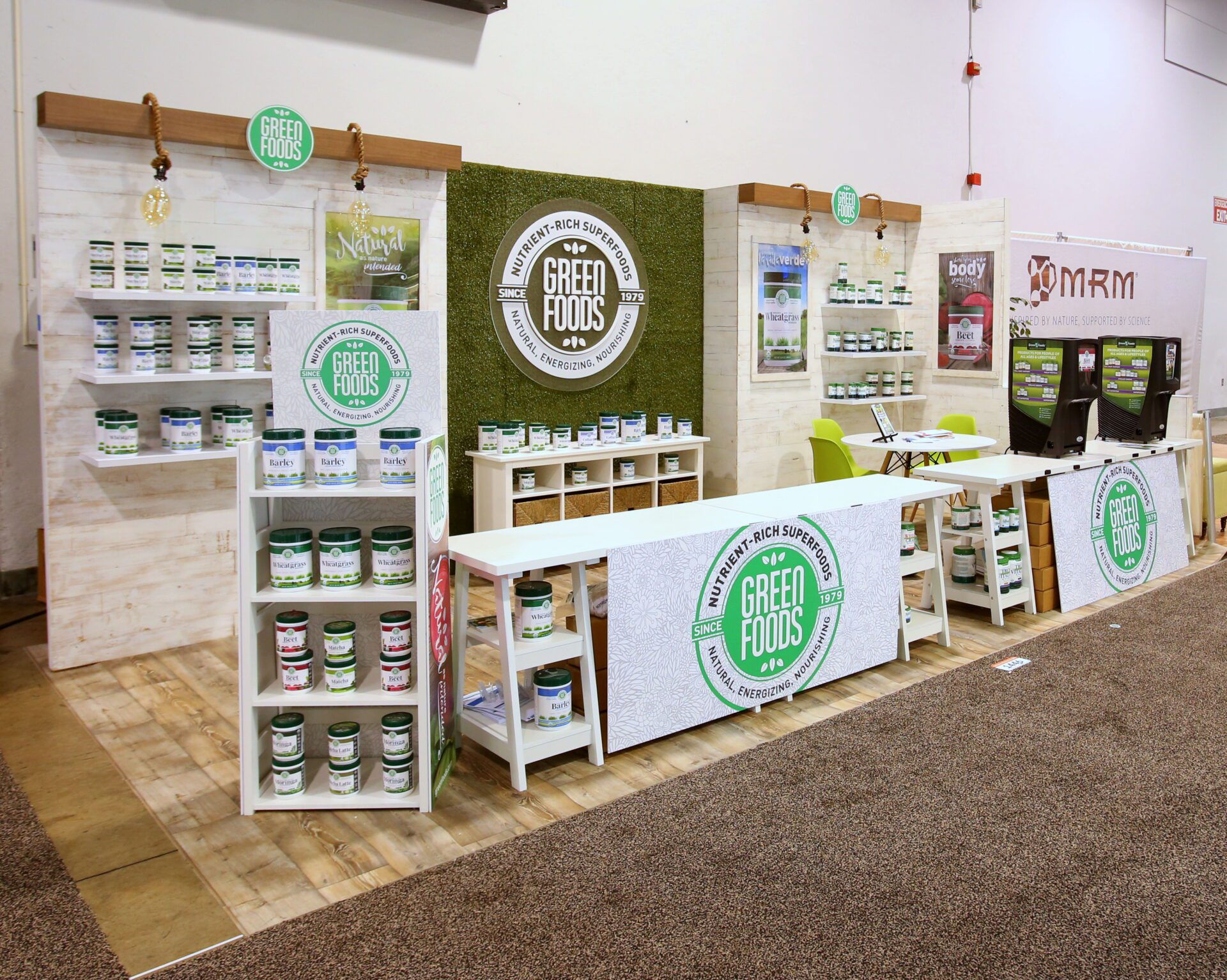 A trade show booth showcasing Green Foods products. The display includes multiple shelves filled with various Green Foods supplements, branded signs, and banners. There is a counter with promotional material and chairs for visitors. A sign for MRM is visible in the background.