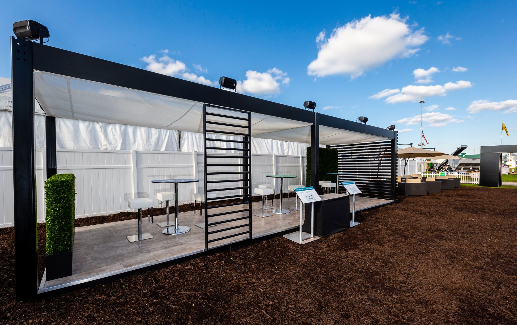 An outdoor seating area with modern black-framed canopies featuring clear roofing and partial white side coverings. Inside, there are high tables with bar stools. Potted plants are positioned near the entrance, and a flag and blue sky with clouds are visible in the background.