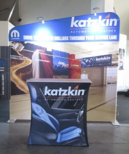 A trade show booth features a display for Katzkin Automotive Leather. A large backdrop shows the Katzkin logo and images of car interiors. A table in front showcases a Katzkin-branded cover with an image of blue leather car seats. There are also banners for various car brands in the background.