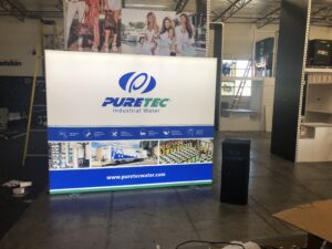 A trade show booth for PURETEC Industrial Water is displayed with a large, illuminated backdrop featuring the company logo, website, and images of their services. The booth area includes a tall, branded stand and other exhibition setups in the surrounding space.