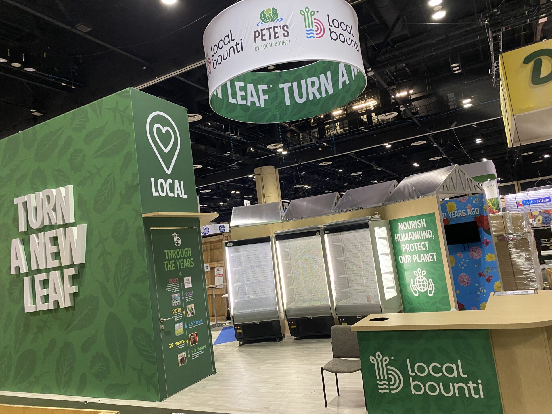 A trade show booth with green walls featuring "Turn a New Leaf" and "Local Bounti" branding. There are informational displays, a seating area with chairs, and a canopy overhead displaying the "Pete's" and "Local Bounti" logos.