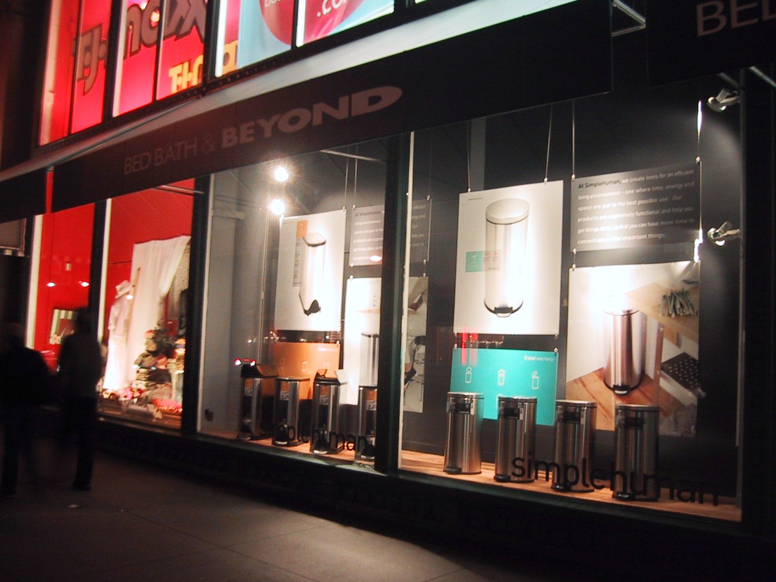 A Bed Bath & Beyond store window display at night showcases various stainless steel trash cans. Large posters of the products hang above, and the store's name and logo are visible on the storefront. The window is illuminated, while people walk by on the sidewalk.