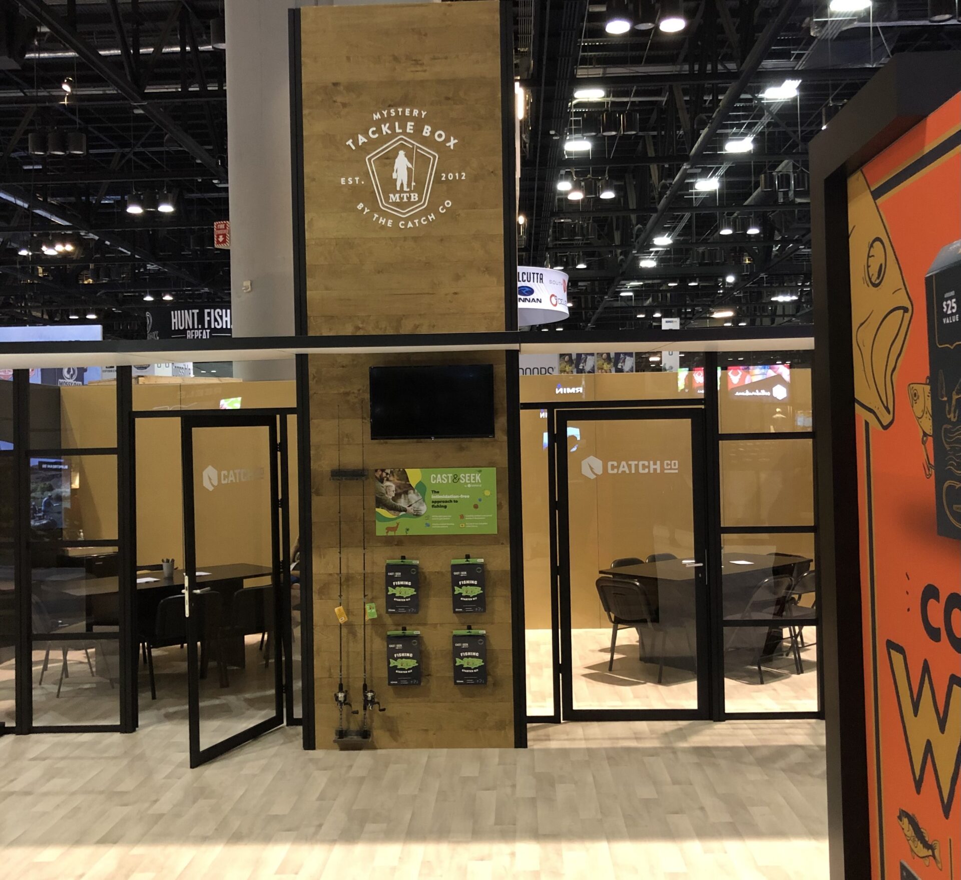 A convention booth featuring Mystery Tackle Box and Catch Co. branding. The booth has wood and glass paneling, with the Mystery Tackle Box logo prominently displayed. There are posters and promotional materials on the walls, and chairs and tables inside the booth.