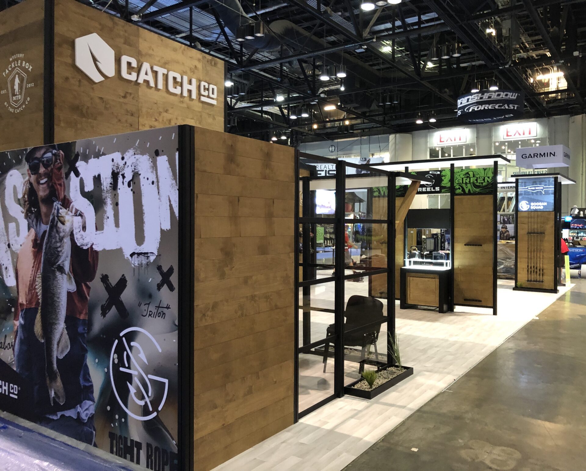 A trade show booth displays the Catch Co brand. It features a section with wooden panels, a large photo of a person holding a fish, and various fishing-related products. Other exhibitors and more displays are seen in the background.