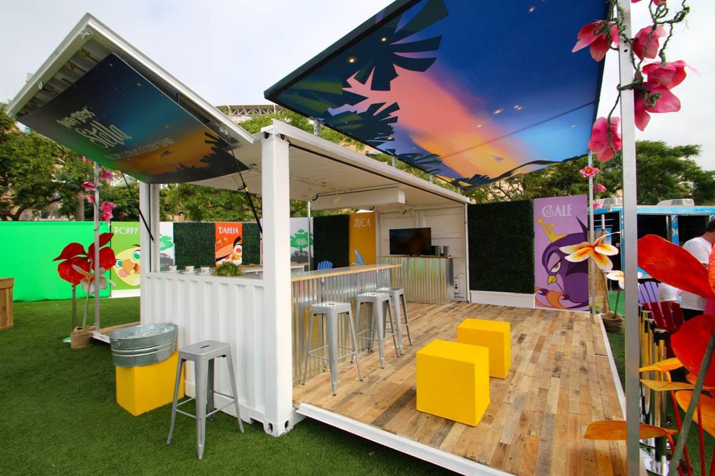 A bright and colorful outdoor pop-up lounge featuring an open-sided container structure with a grass canopy. Inside, there are yellow cube seats and silver bar stools along a corrugated metal counter. Surrounding the lounge are vibrant posters and pink floral decorations.