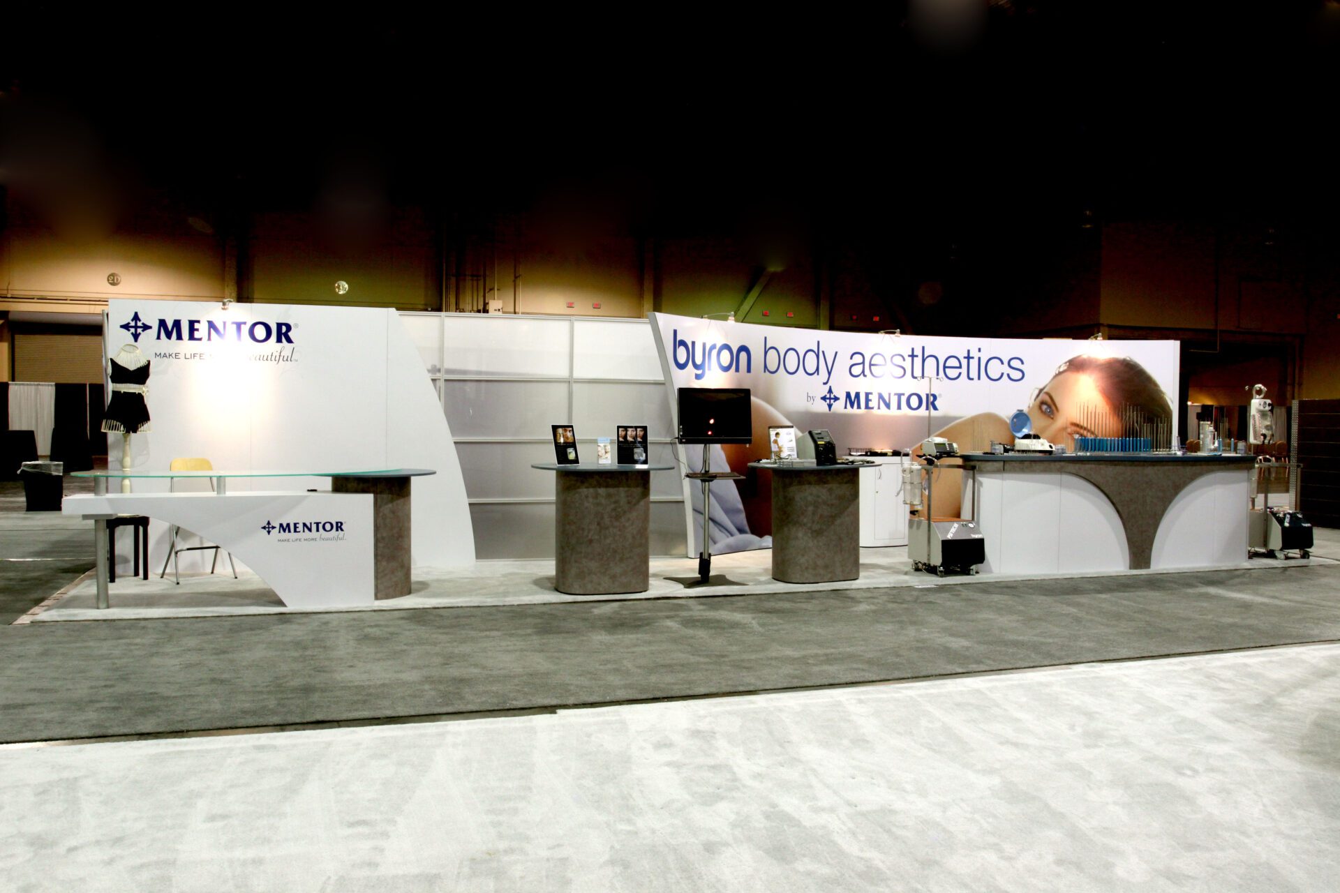 A trade show booth displays the name "Byron Body Aesthetics" and "Mentor" with modern, sleek counters and promotional materials. The setup includes product displays, branding banners, and a backdrop image of a woman's face. The floor is covered with grey carpet.