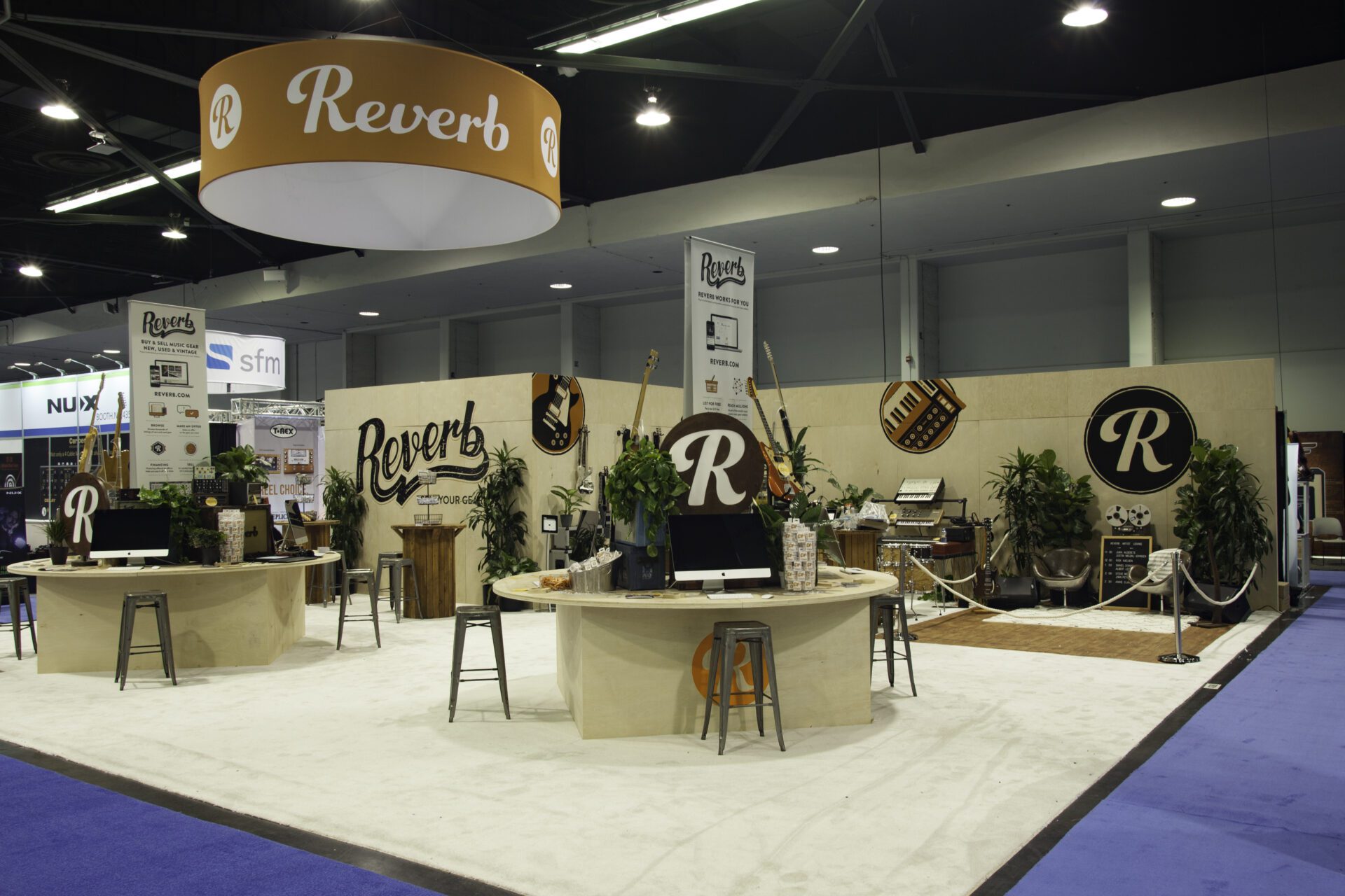 A trade show booth with "Reverb" branding on signs, banners, and displays. Multiple round tables with stools, computers, and potted plants. Musical instruments and audio equipment are displayed in the background. The floor is carpeted in white and blue.