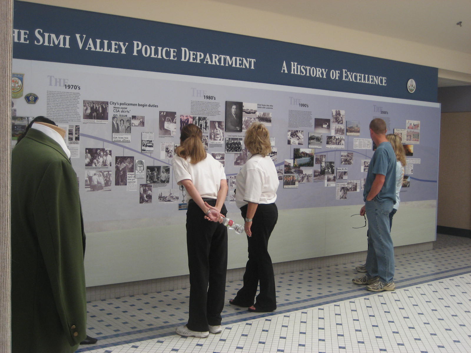 Four people stand in front of a large display board titled "The Simi Valley Police Department: A History of Excellence." The board features various photographs and text detailing the police department's history from different decades.