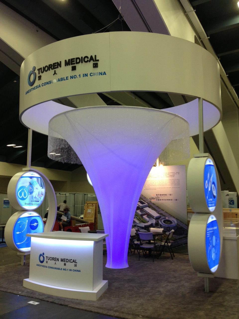 An exhibition booth for Tuoren Medical featuring anesthesia consumables. The booth has a central blue-lit, funnel-shaped structure, a counter with the company's name, and information displays on either side. A banner above reads "Anesthesia Consumables No. 1 in China.