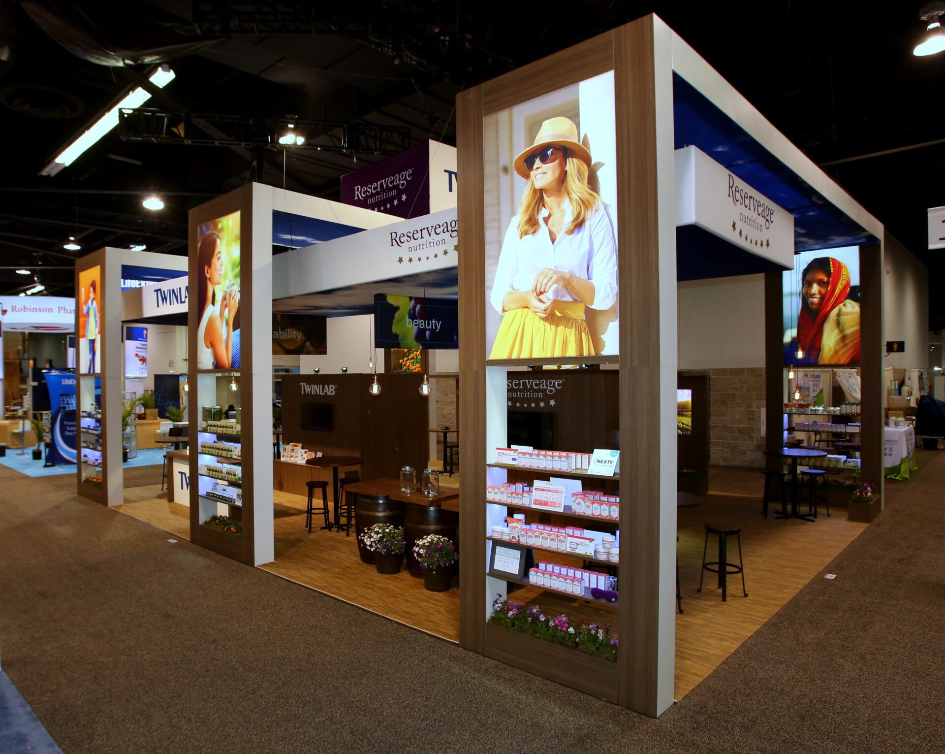 A vibrant exhibition booth showcasing Reserveage products at a trade show. The booth features large images of people, neatly arranged product displays, and seating areas with small tables and chairs. The design has a wood and white color scheme with modern lighting.