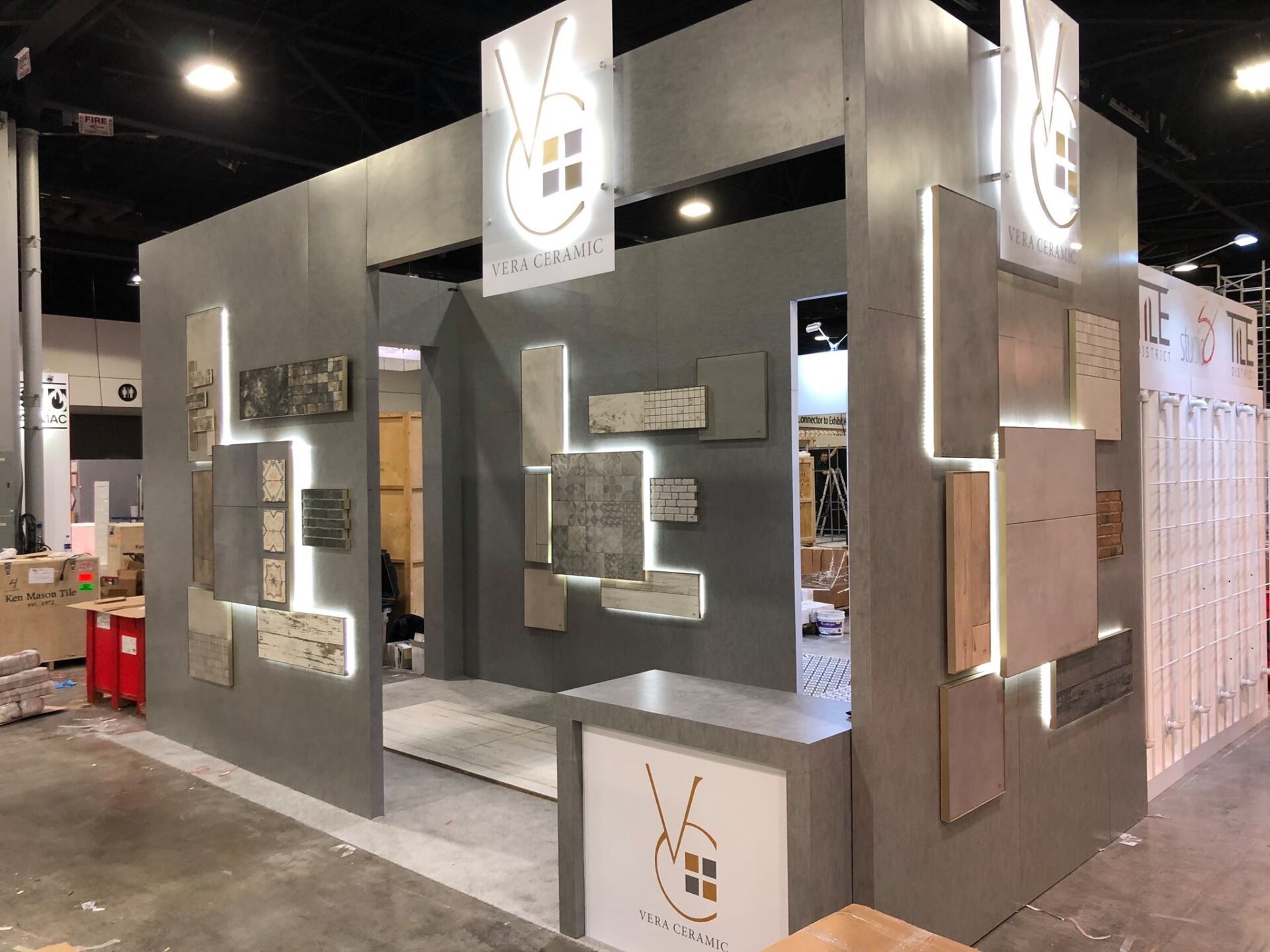 An exhibition booth for Vera Ceramic is displayed at a trade show. The booth features various tile samples arranged on walls with illuminated backlighting. The company's logo is prominently displayed at the top of two corners of the structure.