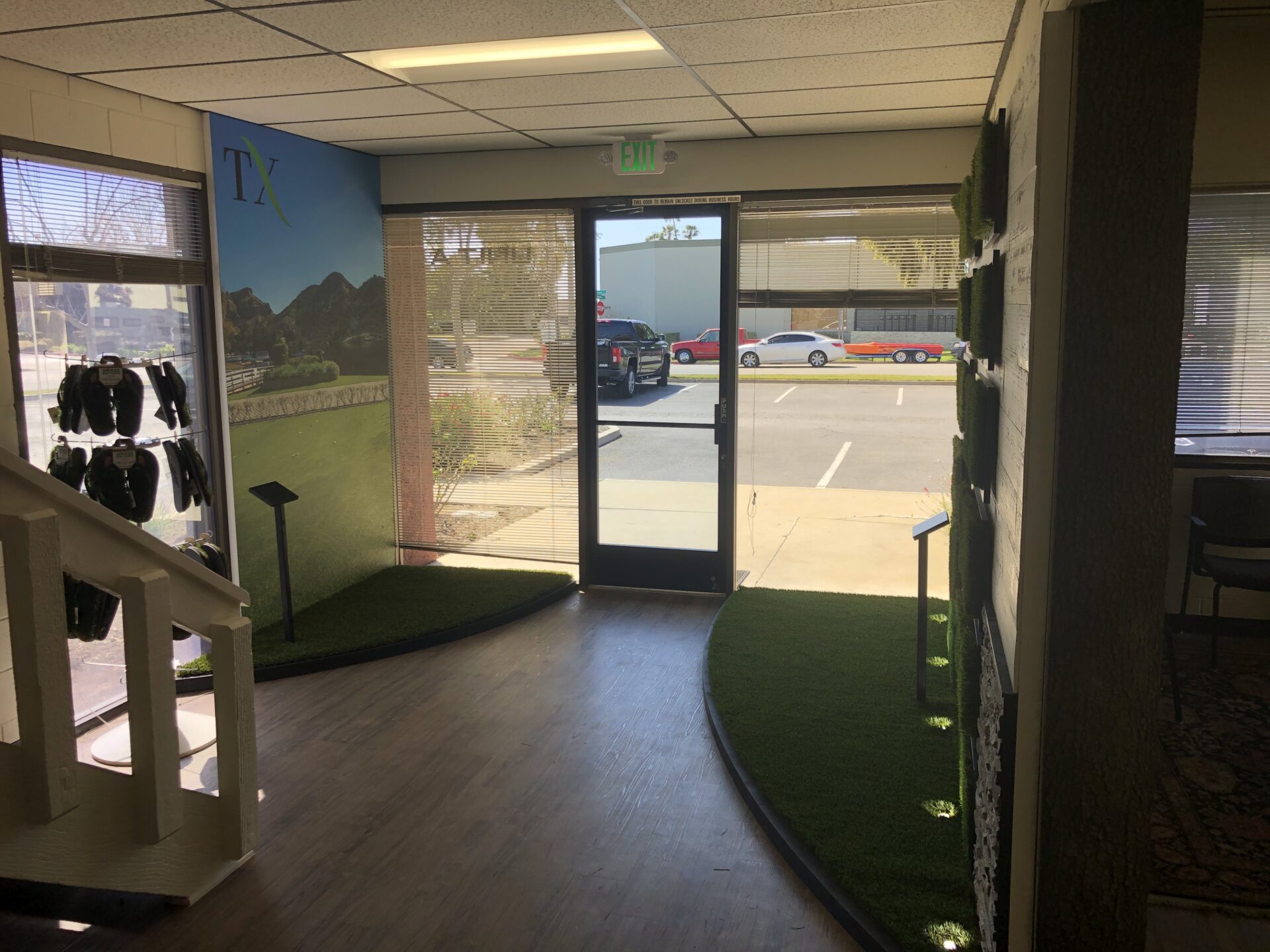 A sunlit entrance of an office building with a glass door and large windows. The walls display green and black products, possibly sports gear. The floor has a mix of wood and green artificial turf. A staircase on the left leads to an upper floor.