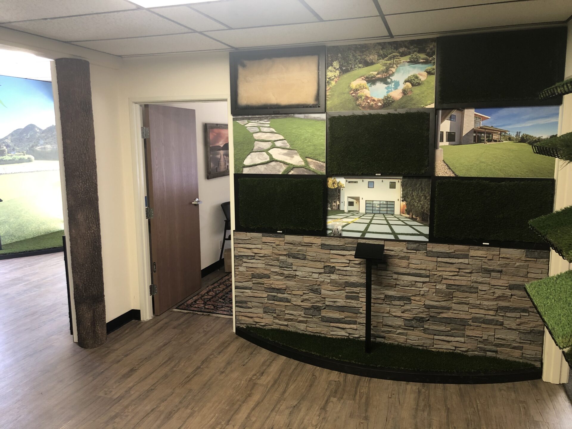 A corner of an interior room showcases various artificial grass patch displays mounted on the walls above a stone veneer base. A door is partially open, leading to another room, and the floor is covered with wood-patterned laminate. Large landscape photos adorn the upper walls.