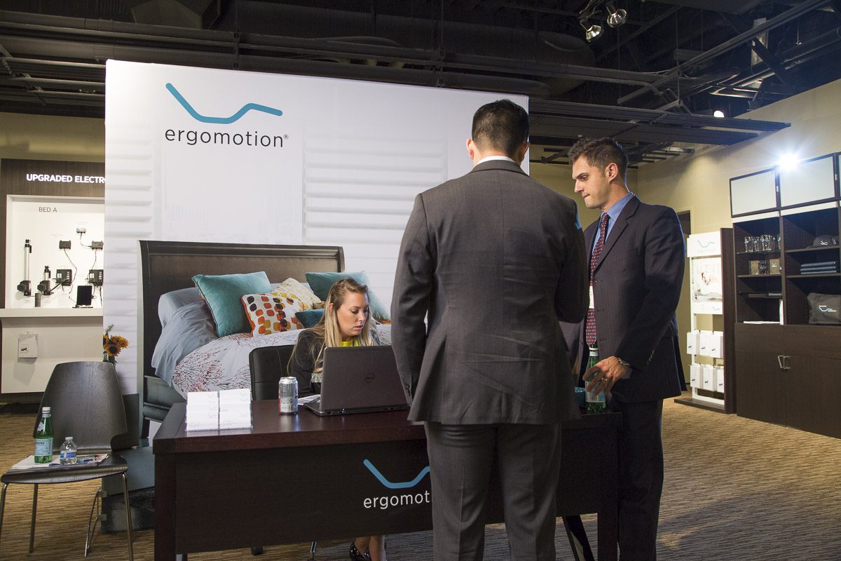 Three individuals in business attire stand around a table with a laptop, engaging in discussion. One person is seated, typing on the laptop. Behind them is a bed display and a banner showing the ergomotion logo. Shelves with various items are visible in the background.