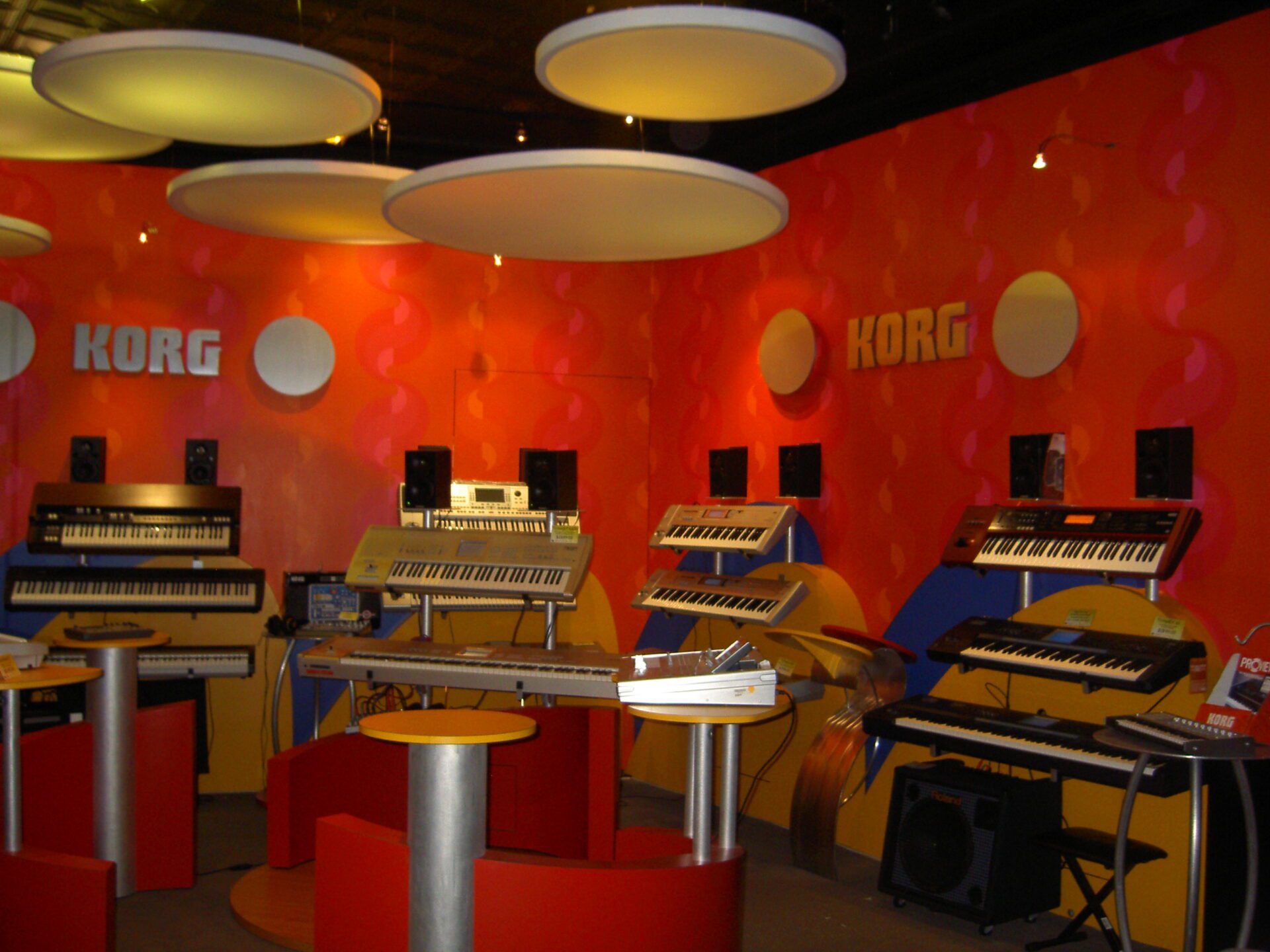 A vibrant showroom with red and orange walls displays multiple Korg keyboards and synthesizers on stands. Circular light fixtures hang from the ceiling, and various audio equipment and speakers are positioned around the room.