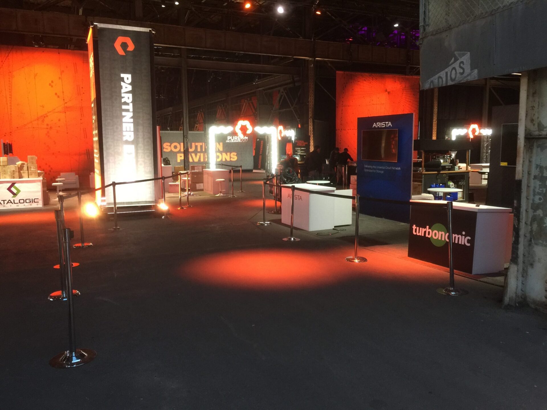 An indoor event setup with various company booths and signage with orange and red lighting. Booths for companies including "Turbomic" and "Arista" are visible. Black stanchions with ropes guide the area, and the space has a warehouse-like industrial feel.