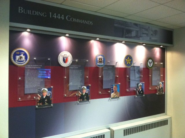 A display wall titled "Building 1444 Commands" showcases emblems and photos of various military personnel, each accompanied by descriptive plaques. The wall has ceiling lights illuminating each section. The background features multiple colors, including shades of red and blue.