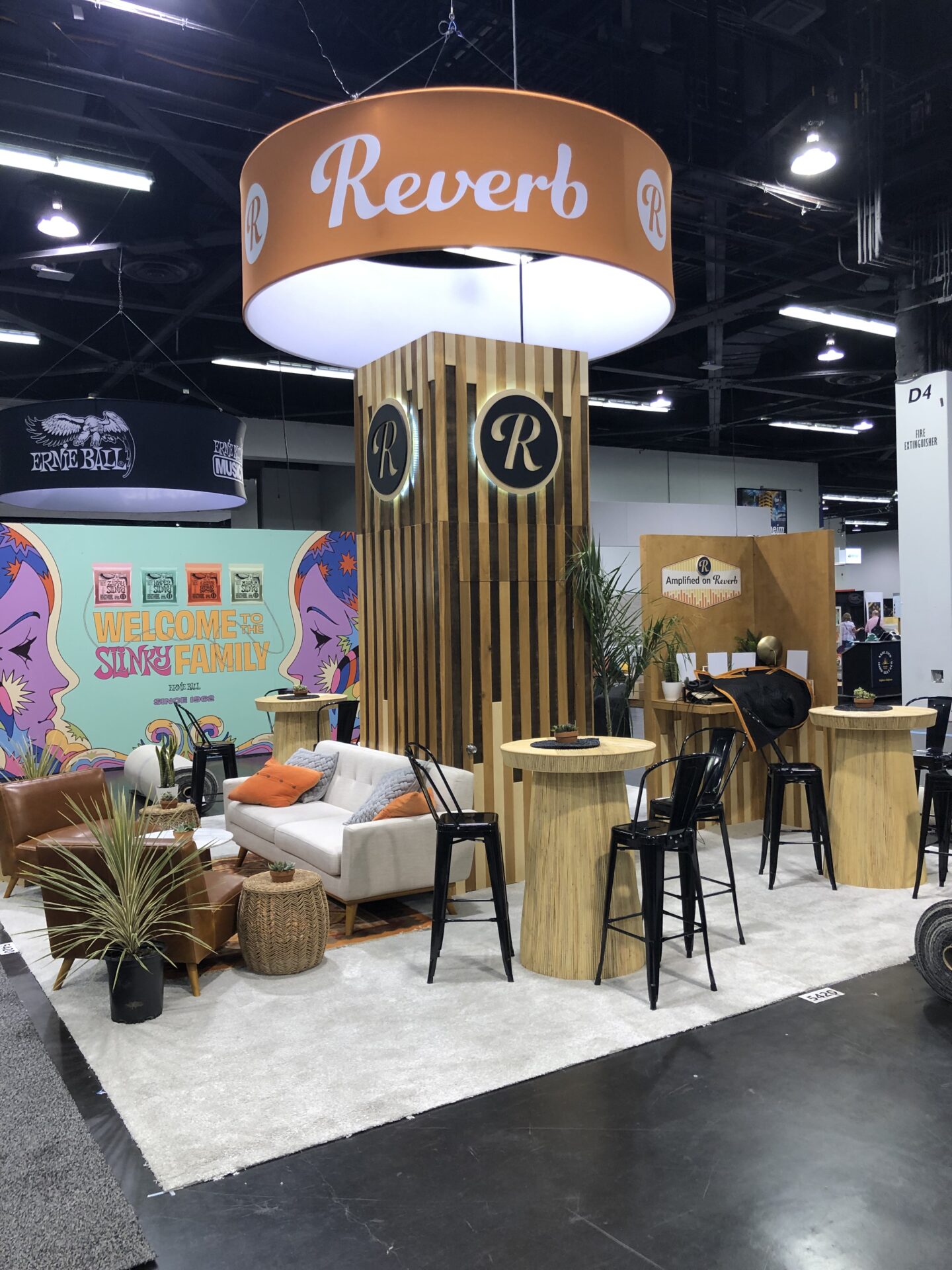 A trade show booth for Reverb with wooden accents, high stools, a couch, and plants. An overhead circular sign with the logo and name "Reverb" is visible. Colorful artwork, including a "Welcome to the Sire Family" sign, is displayed in the background.