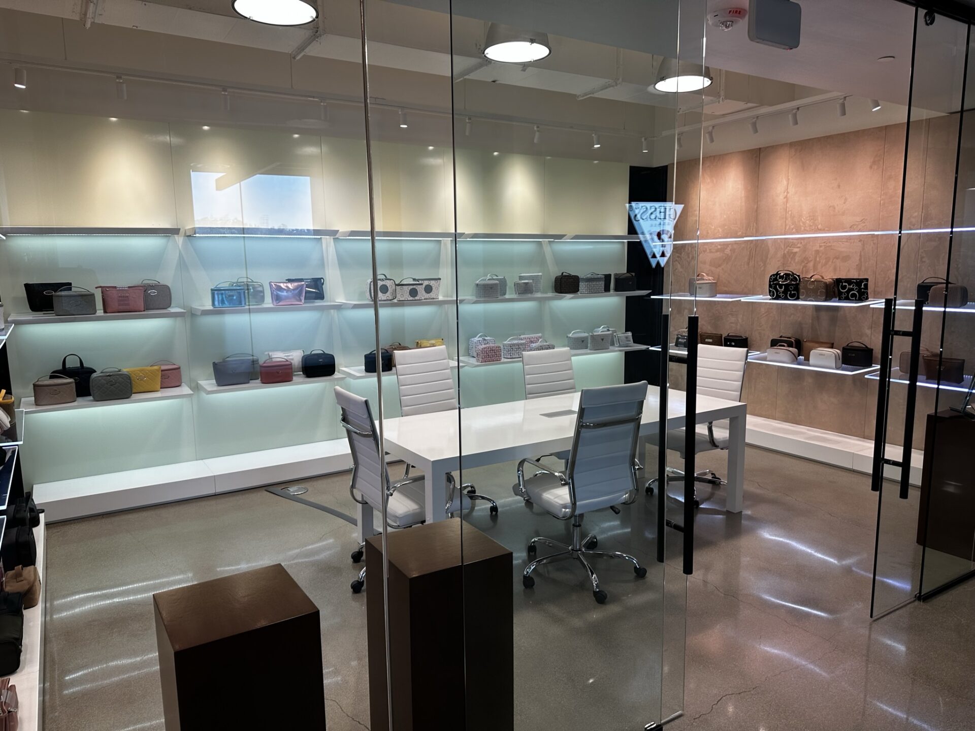 A modern showroom with glass walls featuring designer handbags displayed on minimalist shelves. The room has a sleek white table surrounded by white leather chairs. Recessed lighting illuminates the items on display, creating a luxurious and elegant atmosphere.